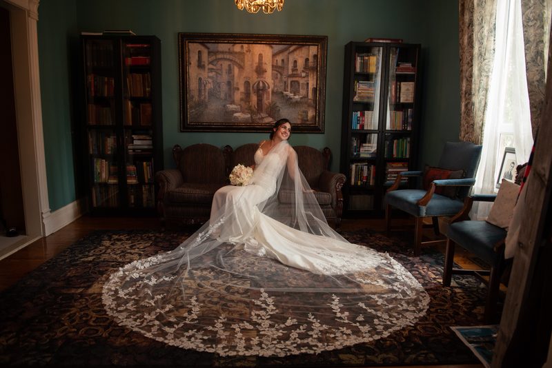 A bride in a white gown and lace veil sits on a vintage sofa in a book-lined room, smiling at the camera.