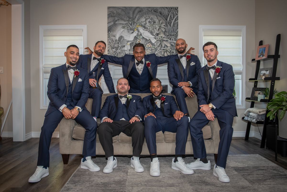Seven men in navy suits pose on and around a beige sofa in a modern living room, relaxed and celebratory.