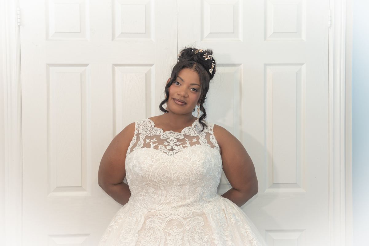 A bride in a white lace gown stands confidently by a door, hands on hips, hair styled with a decorative piece.