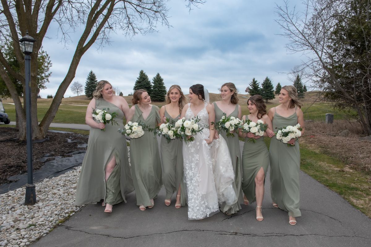 A bride in a white lace gown walks with her smiling bridesmaids in sage green dresses along a paved path.