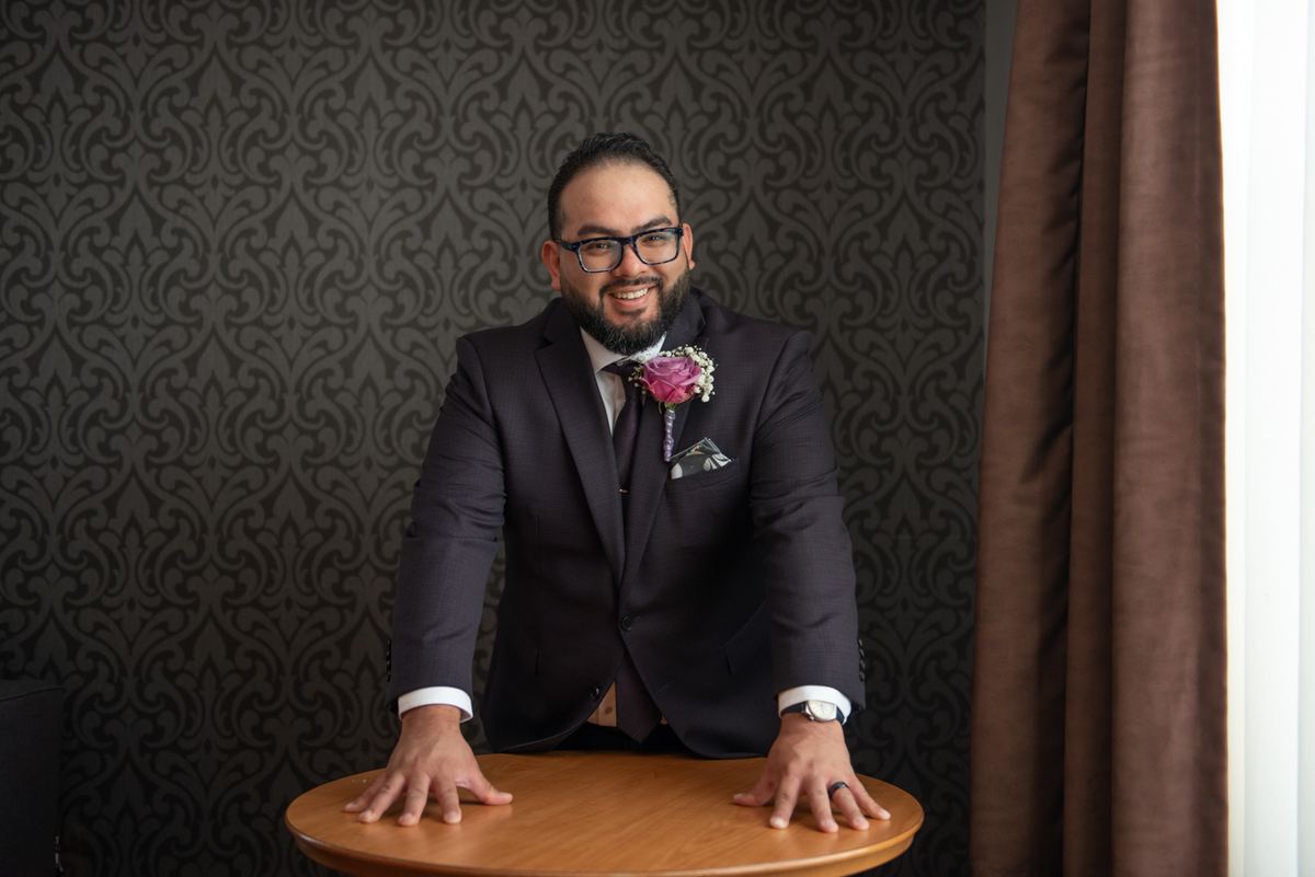 A groom in a dark suit leans forward with both hands on a wooden table, smiling against ornate wallpaper.