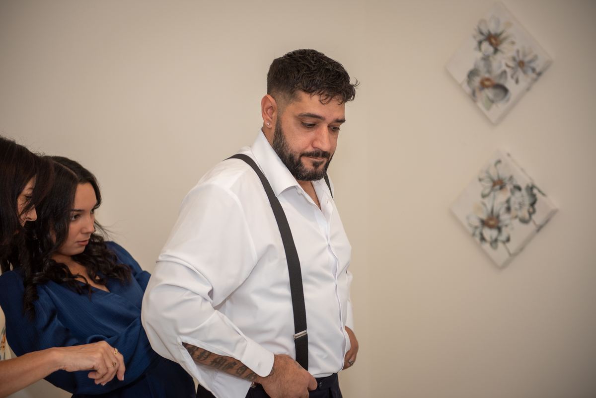A groom in white shirt and black suspenders stands contemplatively as women adjust his attire near floral wall art.