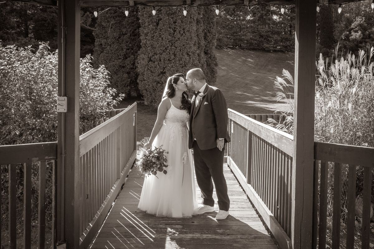 A bride and groom kiss on a wooden bridge surrounded by lush foliage, sunlight casting striped shadows.