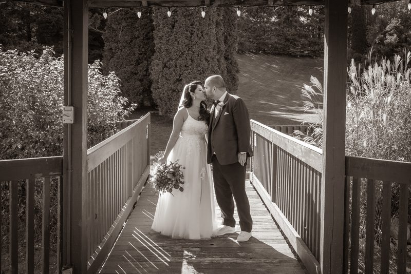 A bride and groom kiss on a wooden bridge surrounded by lush foliage, sunlight casting striped shadows.