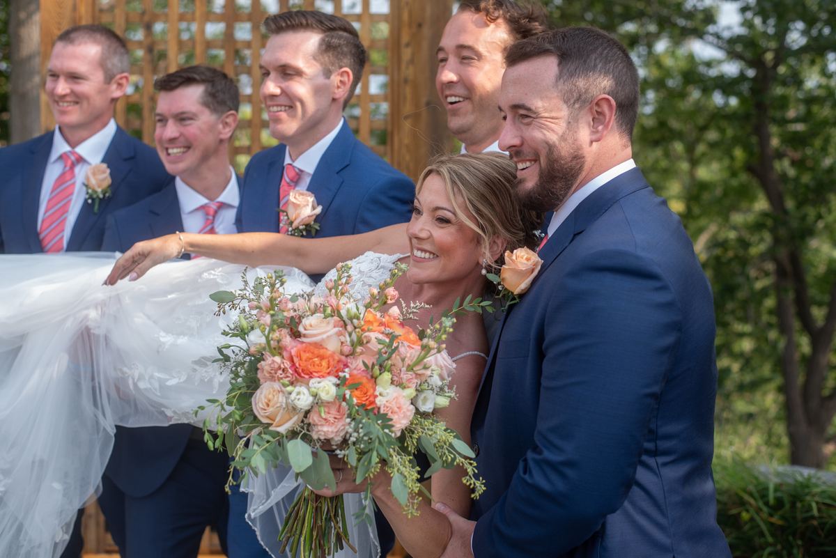 Bride in a white dress smiling with bouquet as groom and groomsman laugh while holding her up outdoors.