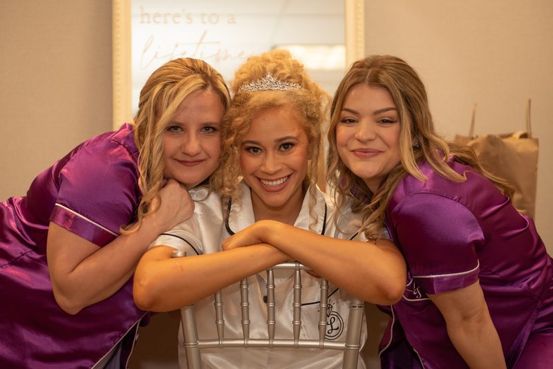 Three bridesmaids pose joyfully around the bride in her bridal suite, arms around her and the chair.