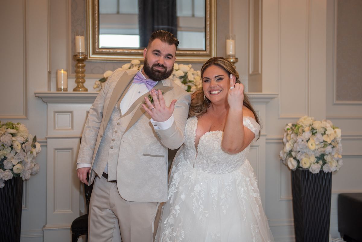 A newlywed couple in formal attire smile joyfully, showing their wedding rings near elegant floral arrangements.