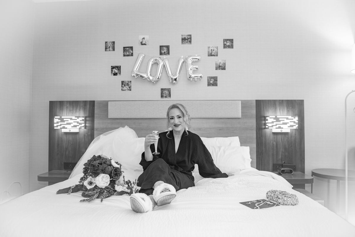 A bride sits happily on a hotel bed with her bouquet and jewelry, surrounded by LOVE balloons and framed photos.