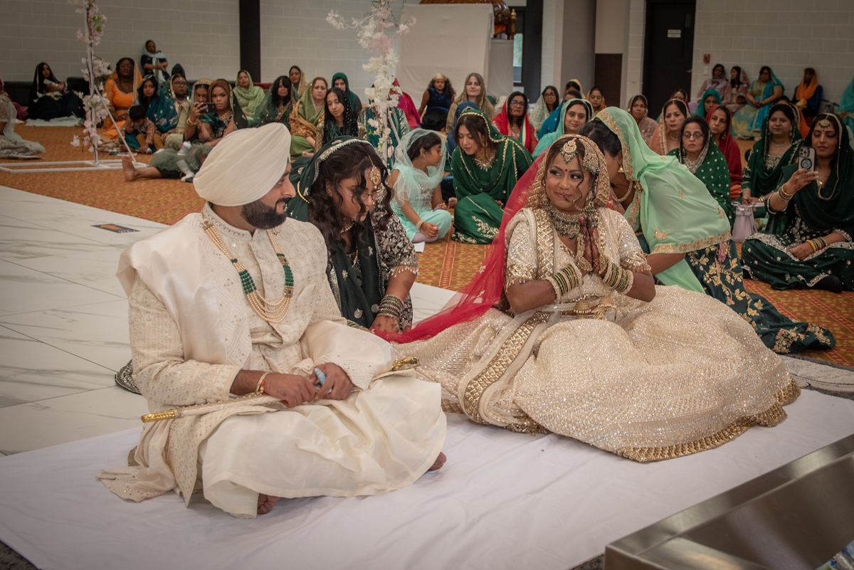 A Sikh groom and bride in traditional attire sit cross-legged with clasped hands, surrounded by family.