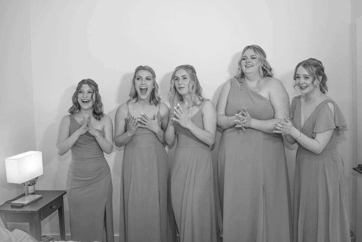Five bridesmaids in matching gowns react with joy, clapping and smiling as one looks amazed at an unseen reveal.