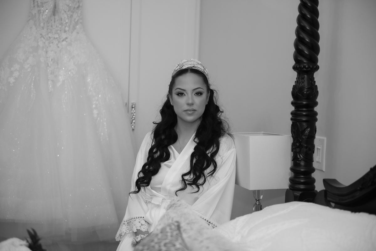 A bride in a white robe and tiara sits thoughtfully in a dimly lit room with her wedding gown hanging nearby.