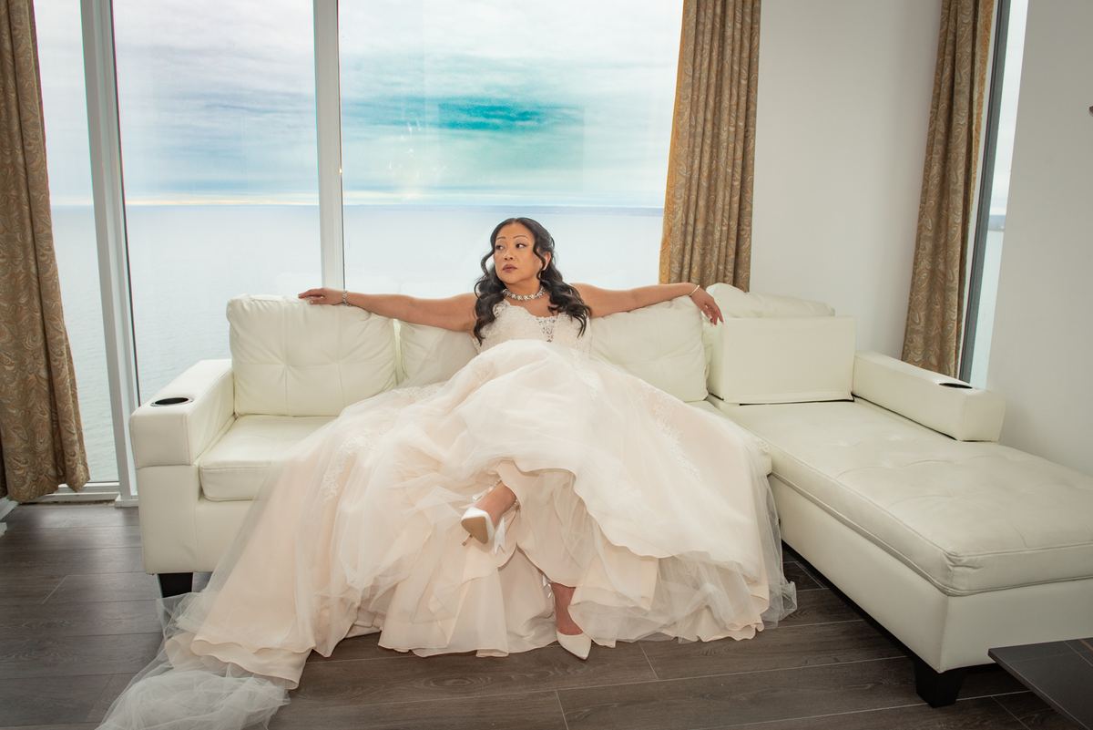 A bride in a white gown rests on a sofa, gazing at the camera with an ocean view through floor-to-ceiling windows behind her.