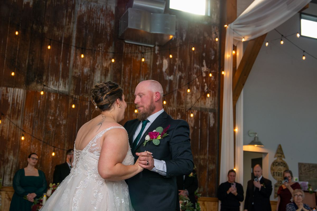 A bride and groom dance closely, gazing at each other in a rustic barn adorned with warm string lights.