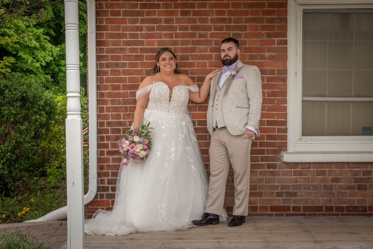 A bride holding pink and white roses and a groom in a beige suit with a lavender bow tie smile on a porch by a brick wall.
