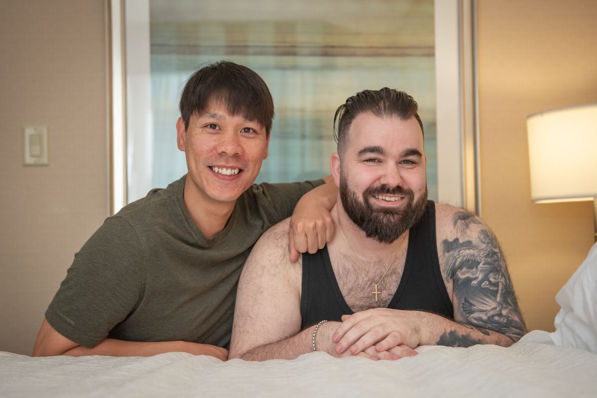 Two men smiling and embracing warmly in a softly lit hotel room, one arm around the other, creating an affectionate scene.