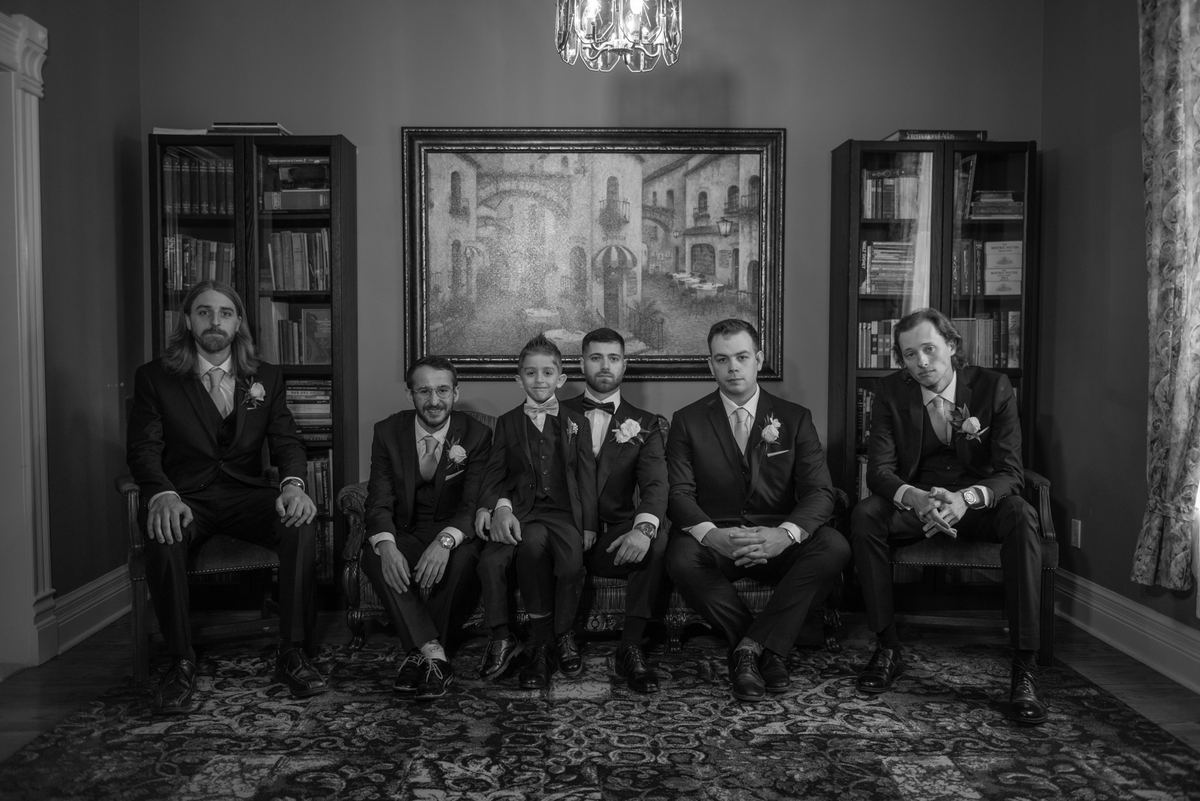 Six formally dressed men and one boy sit in an elegantly decorated room, illuminated by a chandelier.