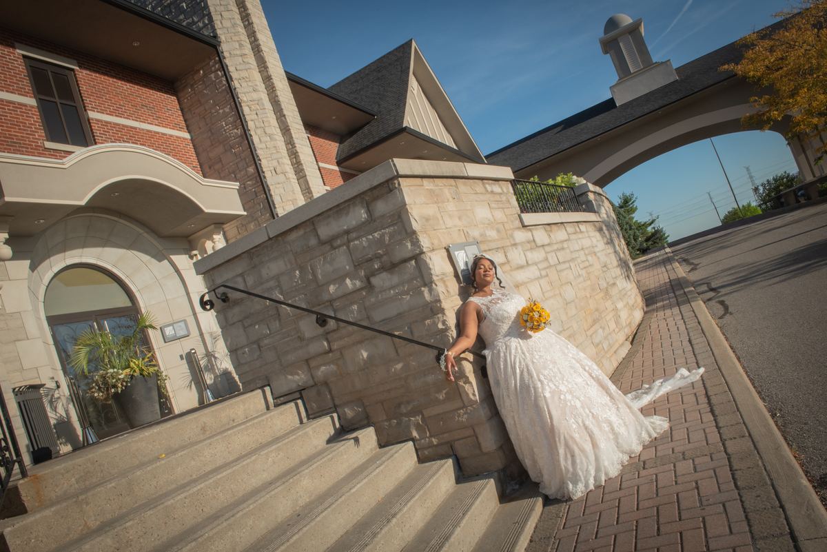 A bride in a white lace gown leans against a stone wall, holding yellow flowers, as sunlight highlights autumn foliage.