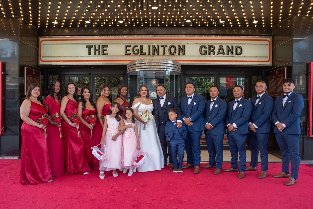 A bride and groom with bridesmaids in red, groomsmen in navy, and two flower girls on a red carpet under string lights.