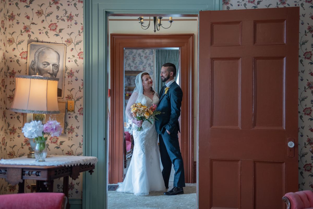 Bride and groom in vintage room share loving glance as soft light from lamp and chandelier enhances moment.