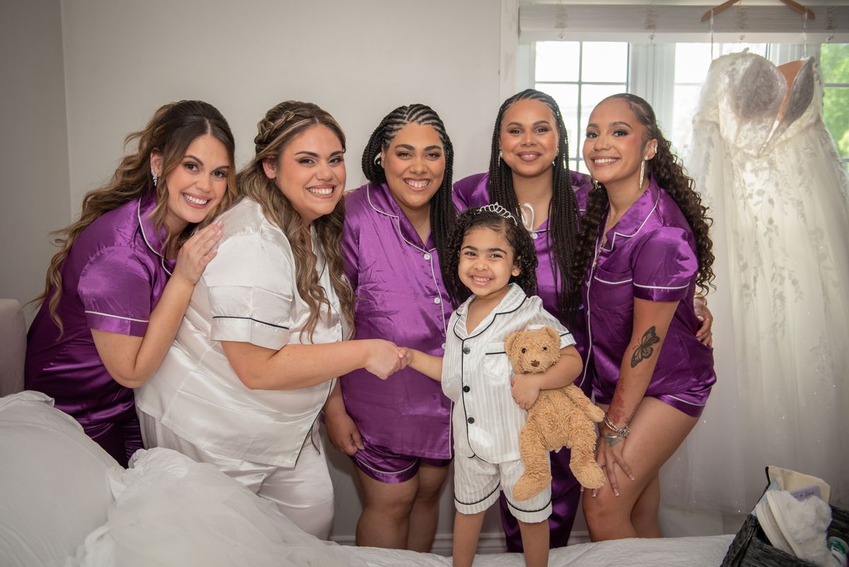 Bride and bridesmaids in satin pajamas gather around a young girl with a teddy bear in a bedroom, smiling.