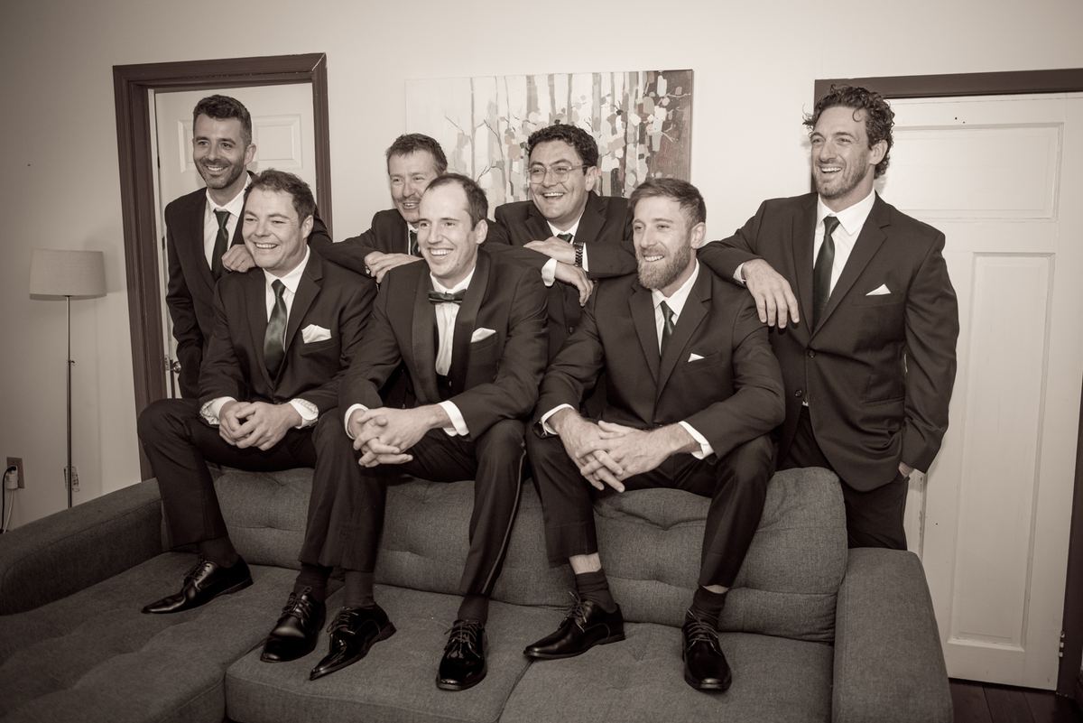 Seven formally dressed men in suits and ties sit and stand together on a couch, smiling and laughing in soft indoor lighting.