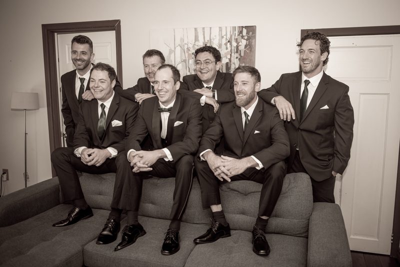 Seven formally dressed men in suits and ties sit and stand together on a couch, smiling and laughing in soft indoor lighting.