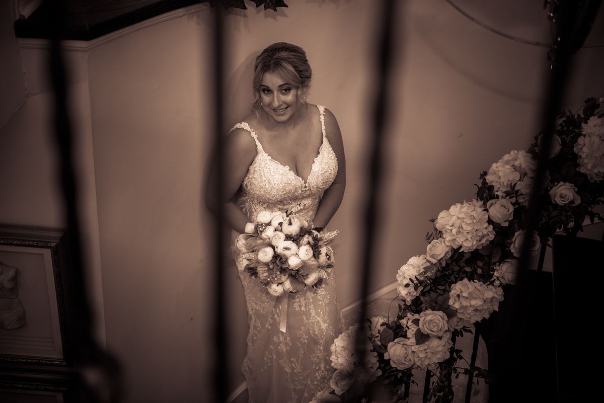 A bride smiles near dark vertical bars, holding a bouquet of white flowers in an intimate, sepia-toned space.