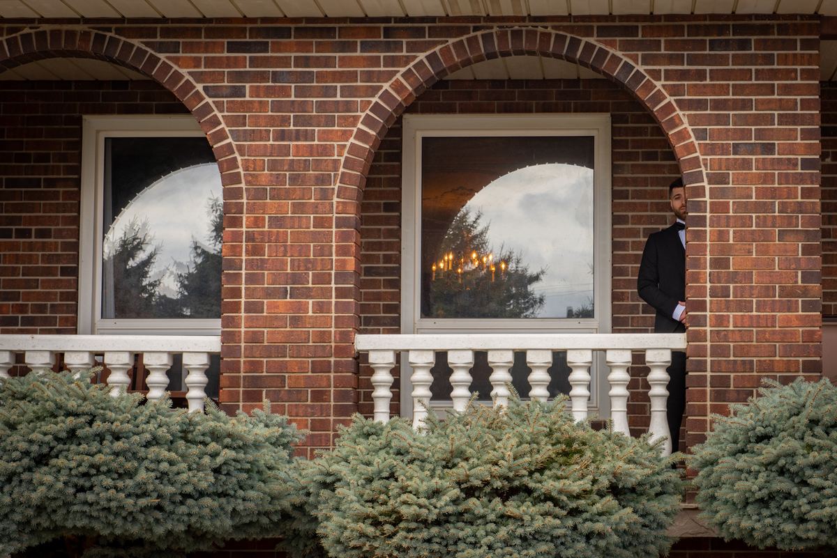 A formally dressed man stands by a brick archway at dusk, with candlelit windows behind him reflecting trees.