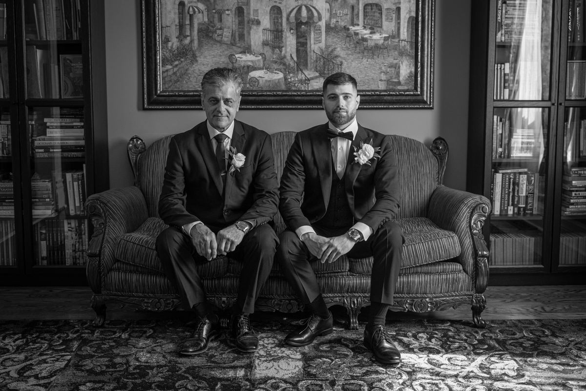 Two formally dressed men sit side-by-side on an ornate sofa in a book-lined room, illuminated by soft light.
