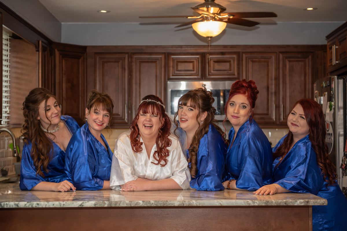 A bride in a white satin robe laughs with her bridesmaids in royal blue satin robes in a cozy kitchen with wooden cabinets.