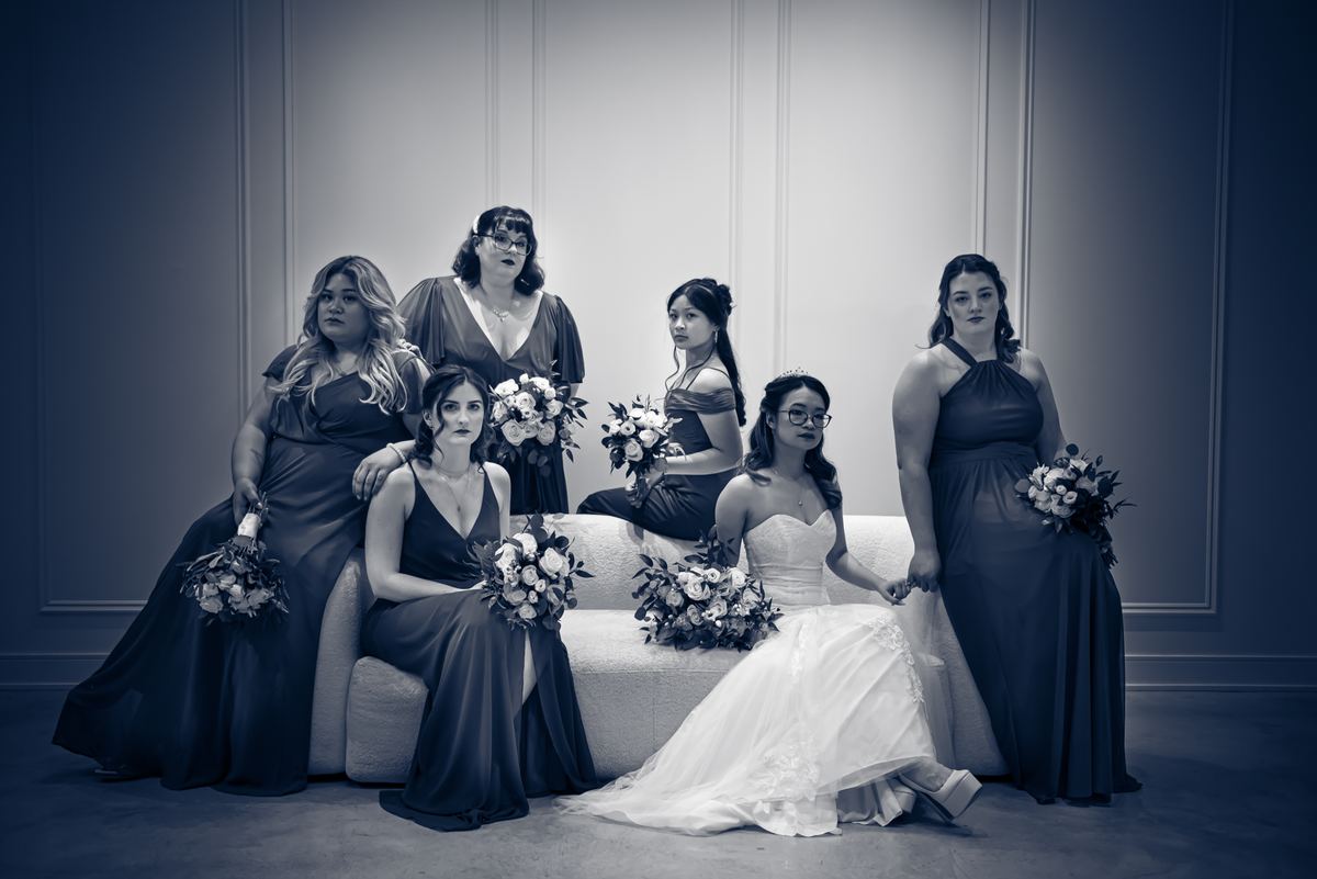A bride in white sits with five bridesmaids in dark gowns on a sofa, illuminated by soft light against a paneled backdrop.