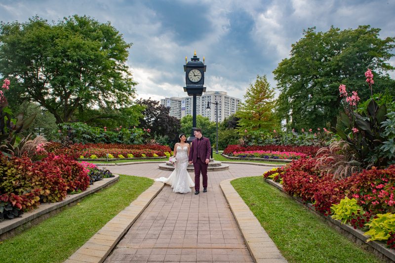 Bride and groom walk hand-in-hand along a garden path framed by flowers and greenery, with a clock tower behind them.