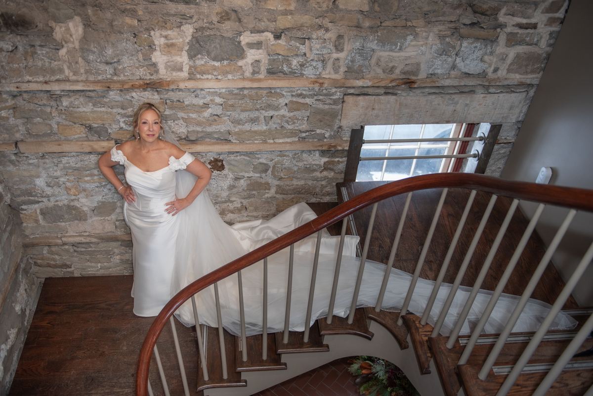 A bride in a white off-the-shoulder gown leans against a rustic stone wall near a window on a wooden staircase.