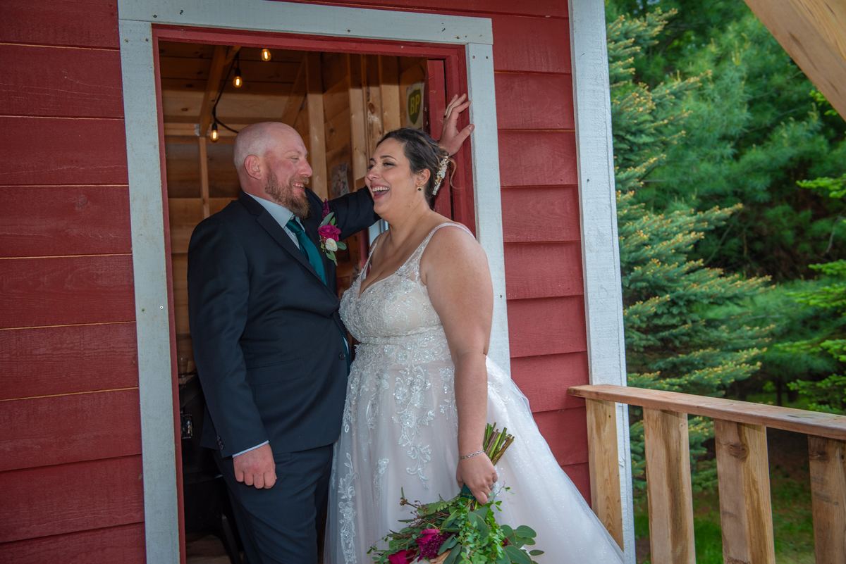A smiling bride and groom laugh together in a doorway surrounded by greenery, illuminated by natural light.