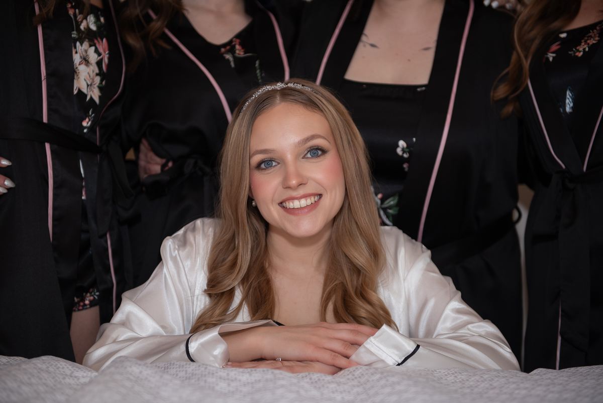 A bride smiles next to bridesmaids in black robes with pink trim, surrounded by soft lighting and satin textures.