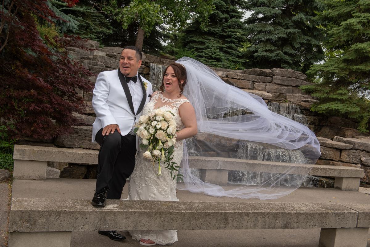 A laughing bride and groom sit on stone steps beside a waterfall, with her veil billowing in the wind.