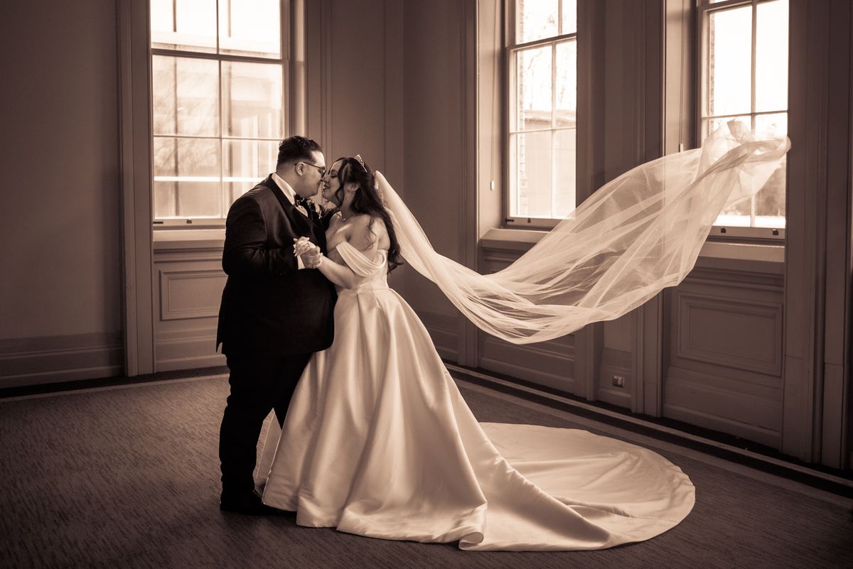 A newlywed couple kisses in a sunlit room, their embrace framed by tall windows casting golden light, veil billowing.