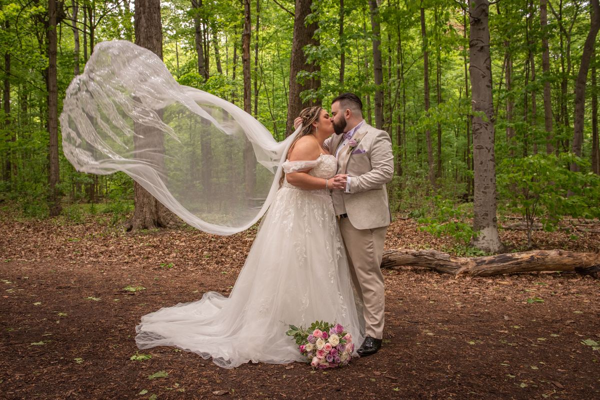 Bride and groom kiss in a sunlit forest, surrounded by trees and her flowing veil dancing in the breeze.