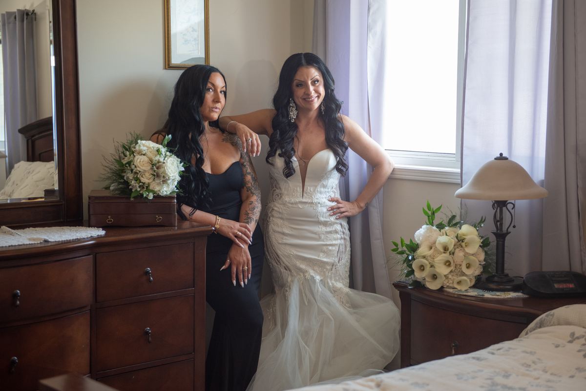 Two women in wedding and black dresses embrace warmly in a softly lit bedroom with floral arrangements.