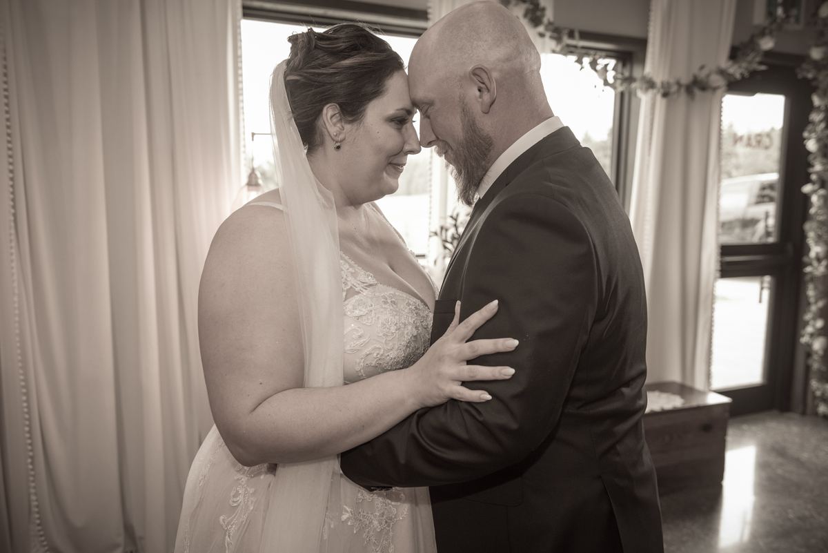 A bride and groom stand close, foreheads touching, in an intimate embrace, illuminated by soft light through windows.