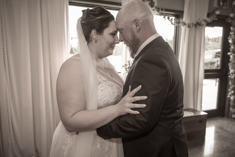 A bride and groom stand close, foreheads touching, in an intimate embrace, illuminated by soft light through windows.