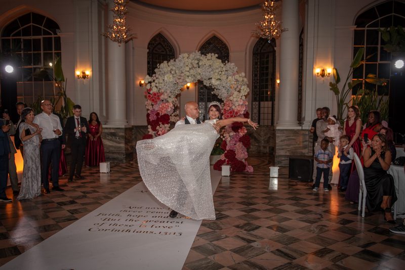 Bride and groom dance joyfully in a grand hall with chandeliers, floral backdrop, and checkered floor, guests watching.