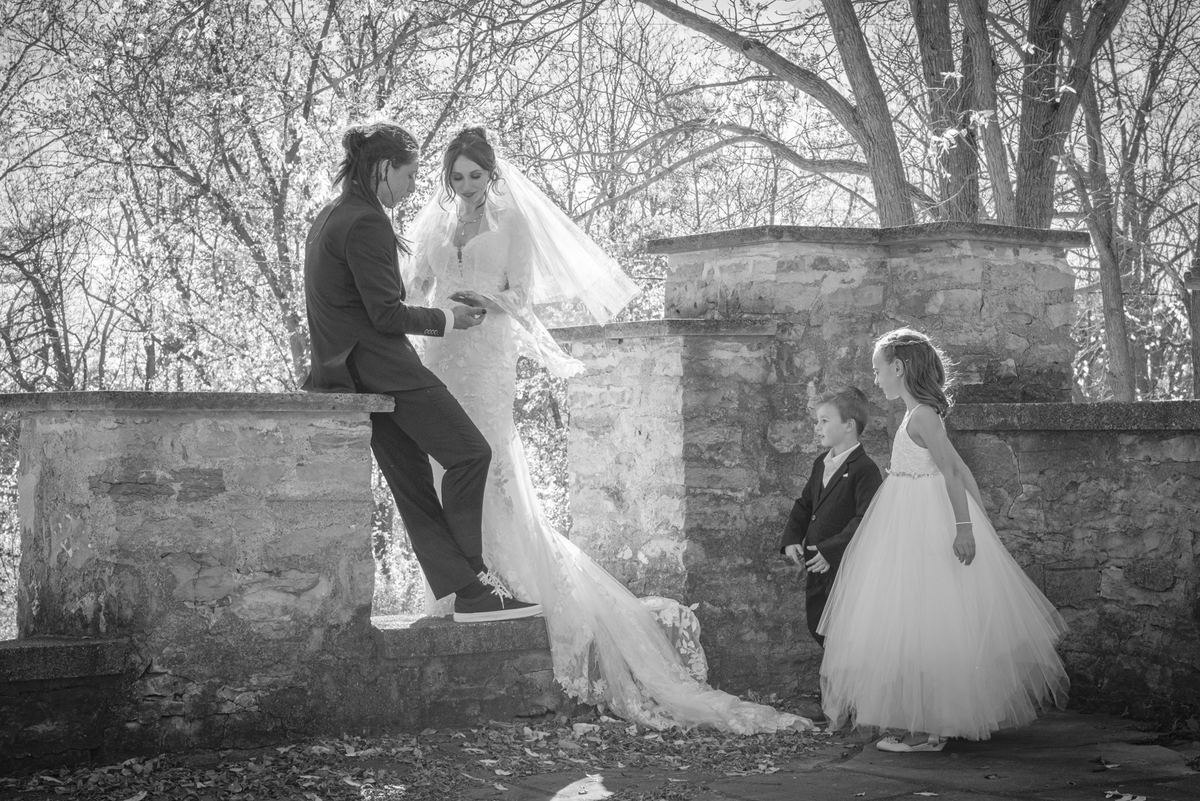 A bride and groom sit on stone steps in a wooded area, signing documents while two children observe.