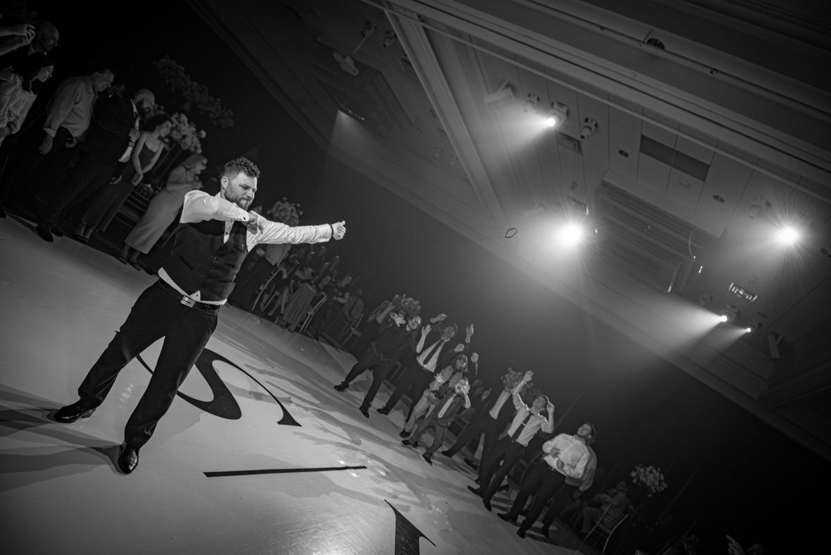 A man in formal attire dances energetically on a glossy floor under bright lights, surrounded by clapping guests.