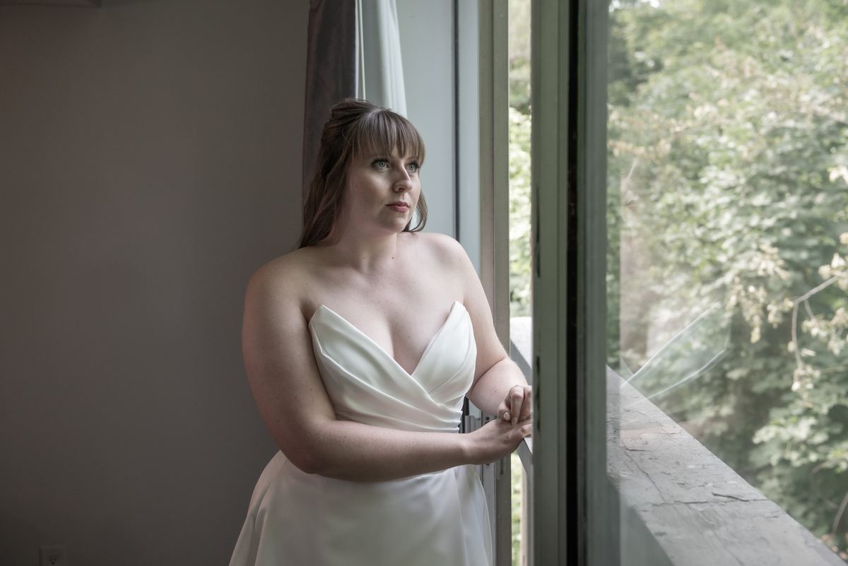 A bride in a white strapless gown stands by a window, gazing outward with her hand resting on the windowsill.