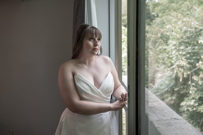 A bride in a white strapless gown stands by a window, gazing outward with her hand resting on the windowsill.