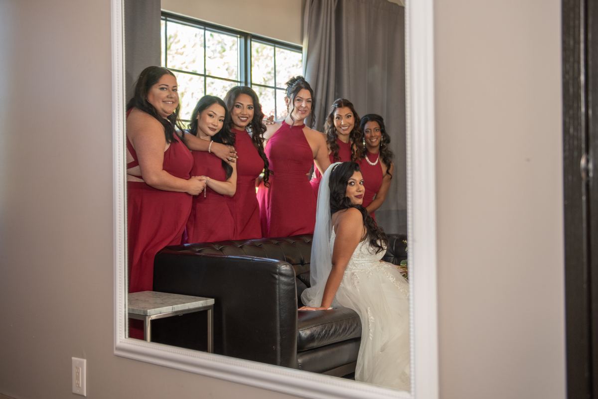 A bride in a white gown sits on a leather couch with her bridesmaids in matching burgundy dresses behind her.