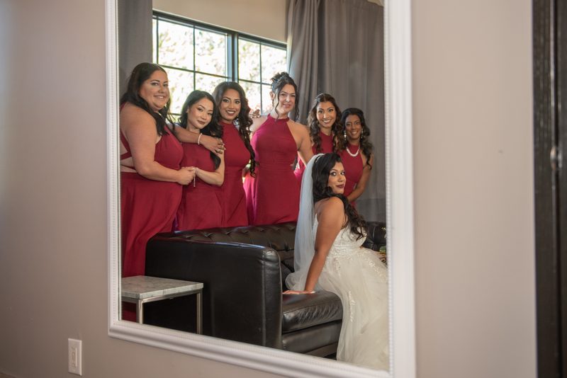 A bride in a white gown sits on a leather couch with her bridesmaids in matching burgundy dresses behind her.