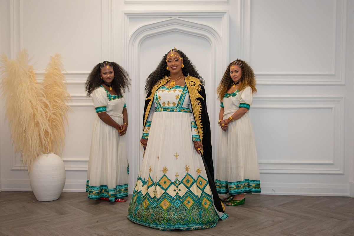 Three women in ornate dresses stand in a minimalist room, with the central figure in a white and teal gown smiling.