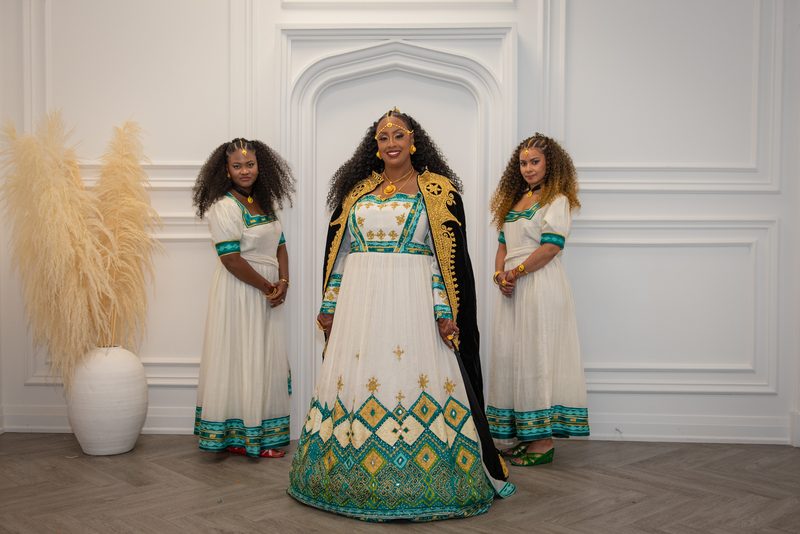 Three women in ornate dresses stand in a minimalist room, with the central figure in a white and teal gown smiling.